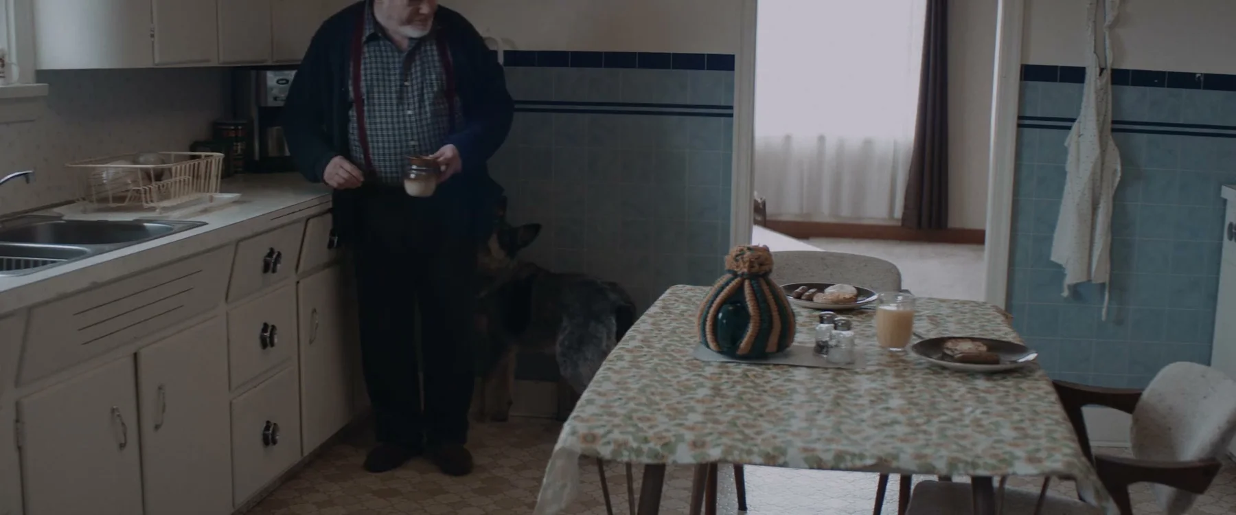 The shopkeeper standing at a small kitchen table with a mug in Mabel
